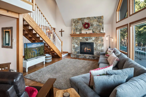 Cozy living room with a stone fireplace, large windows, a staircase, and comfortable seating.